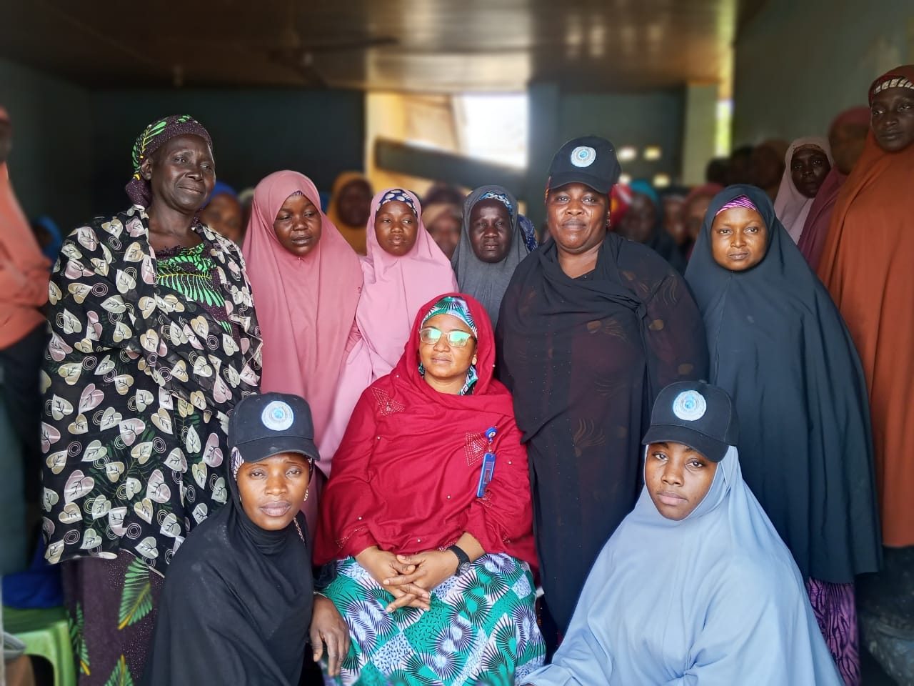 A seated woman in a red shawl surrounded by women in colorful headscarves, two of them wearing caps with the foundation emblem